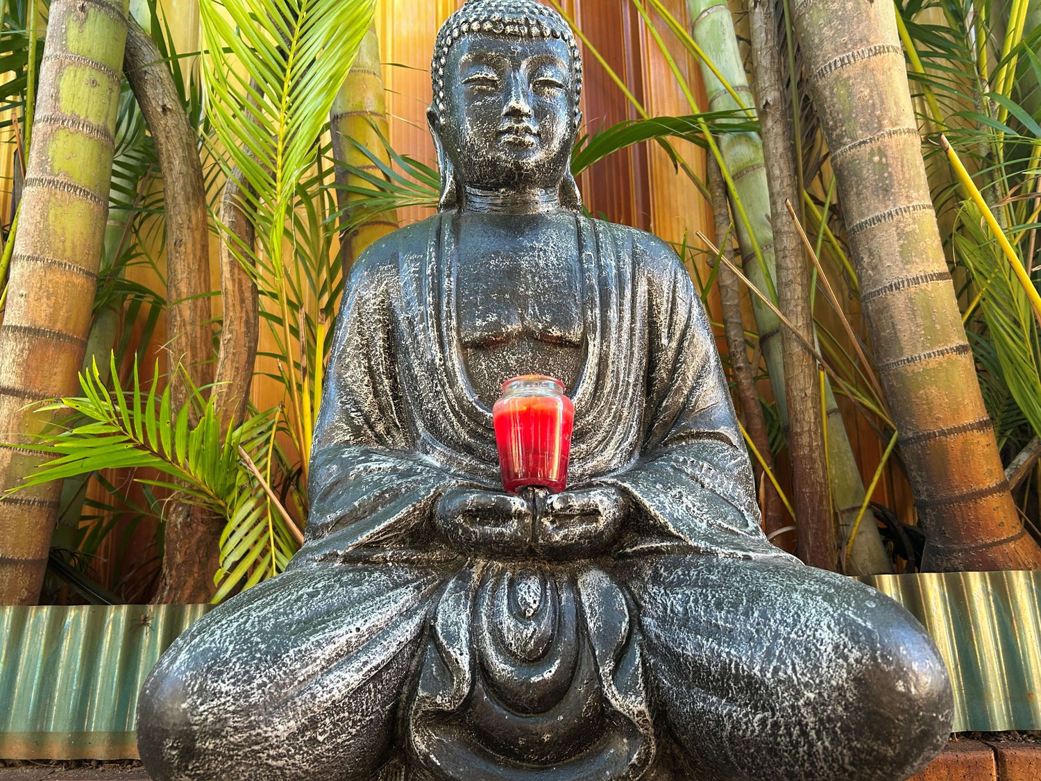 Buddha statue holding a red drink surrounded by green plants, symbolizing peace and mindfulness.