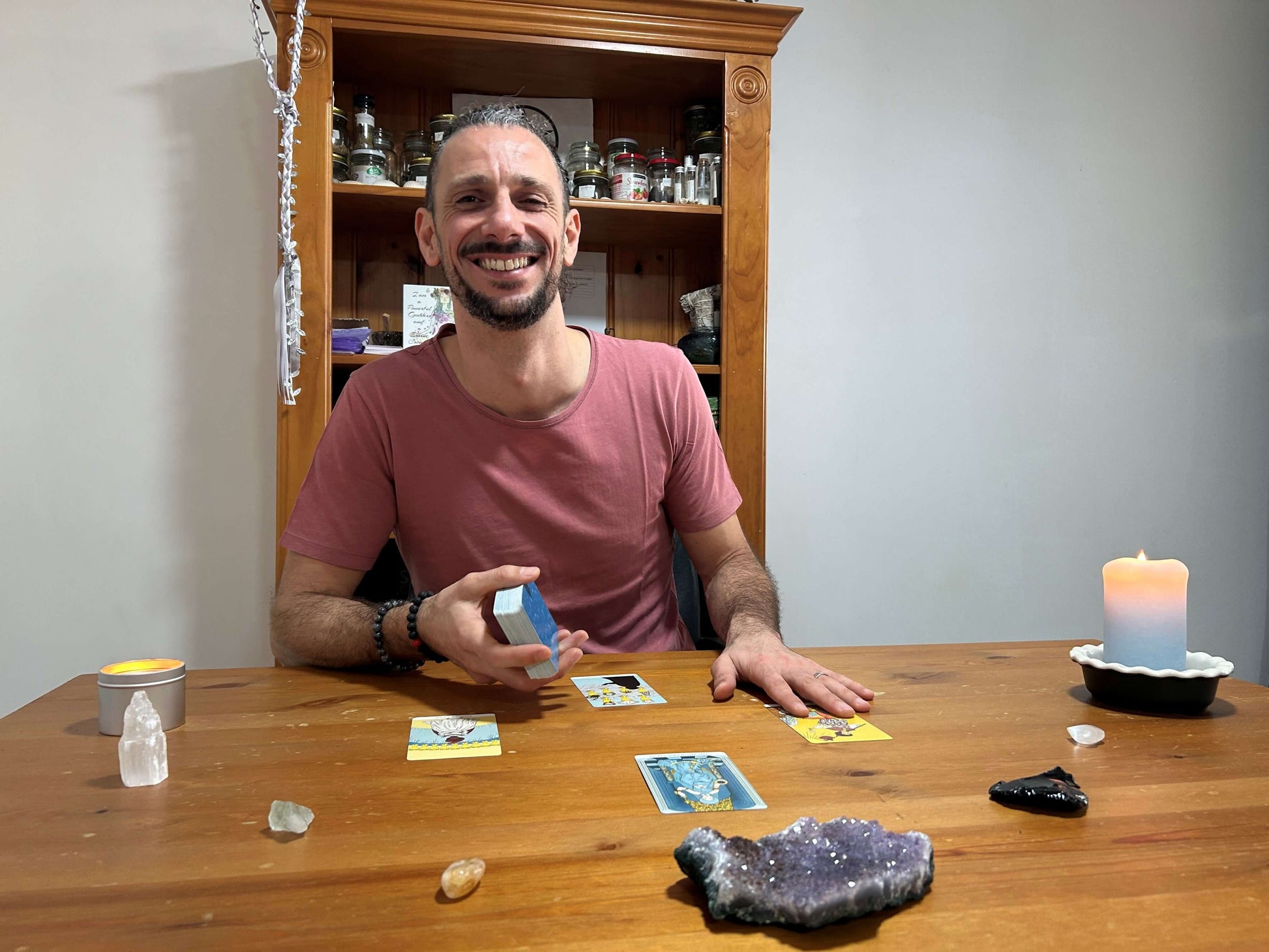 Man sitting at a table with tarot cards, crystals, and candles in a room.